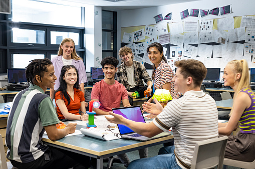 Wide shot of a group of students and their teacher gathered around a table in a technology classroom. They are all wearing casual clothing and are situated in a secondary school in Gateshead, England.