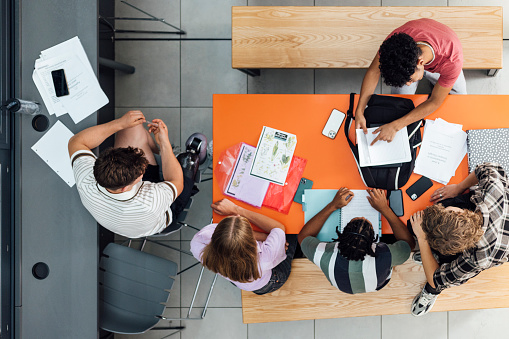 An aerial shot of a group of secondary school students working together in a common room in a school in Gateshead, England. They are all wearing casual clothing and are gathered around worksheets and papers on a table.