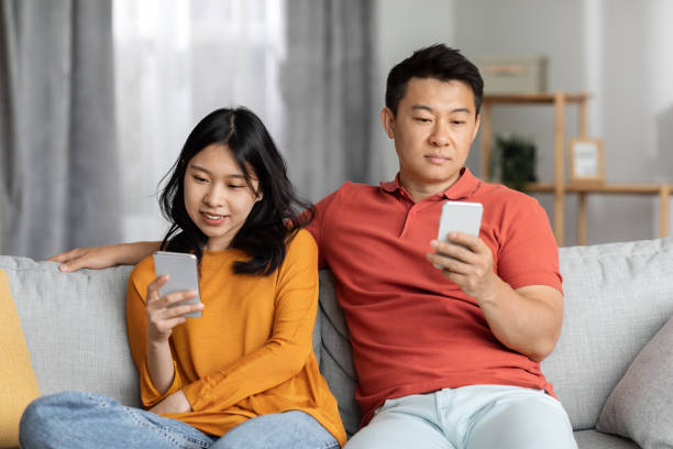 istock Asian couple sitting on couch together, using their smartphones 1469940690