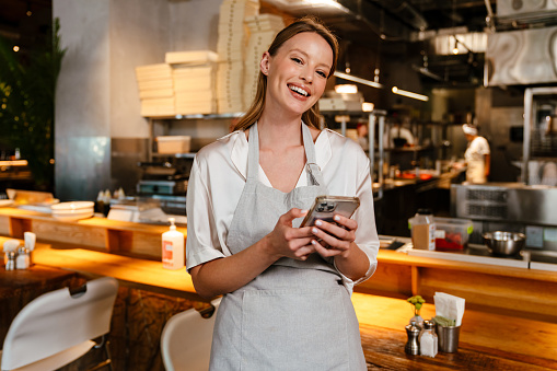 Young blonde woman wearing apron using cellphone while working in cafe