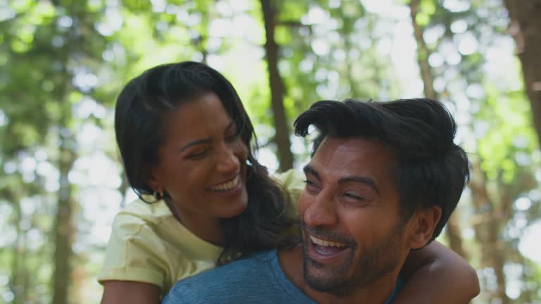 Portrait Of Couple Hiking Or Walking Through Woodland With Man Giving Woman Piggyback