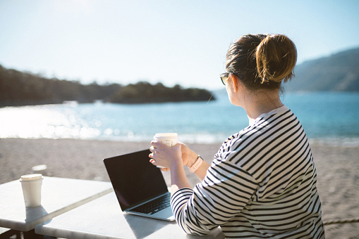 Working outdoors. Mid-aged woman using laptop sitting on the beach and drinking coffee