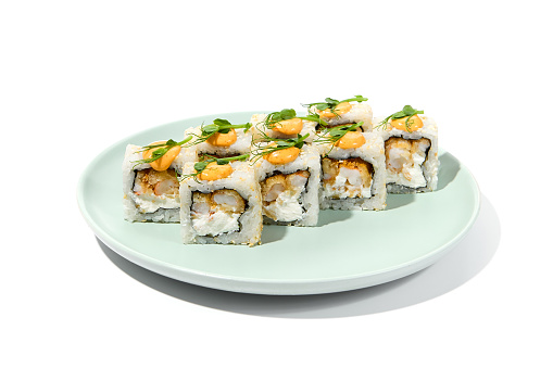 Maki sushi with sesame outside, tempura shrimp inside topped spicy sauce and microgreens. Spicy shrimp maki roll on ceramic plate isolated on white background. Sushi roll with crispy prawn on plate