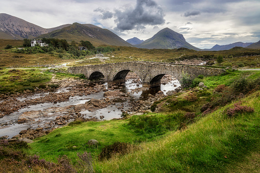 Water flowing under old stone bridge with mountains in background.