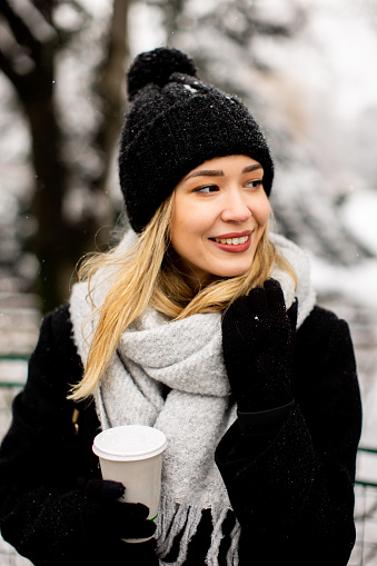 A young woman stands in a winter wonderland, wearing warm clothing and drinking hot coffee to go