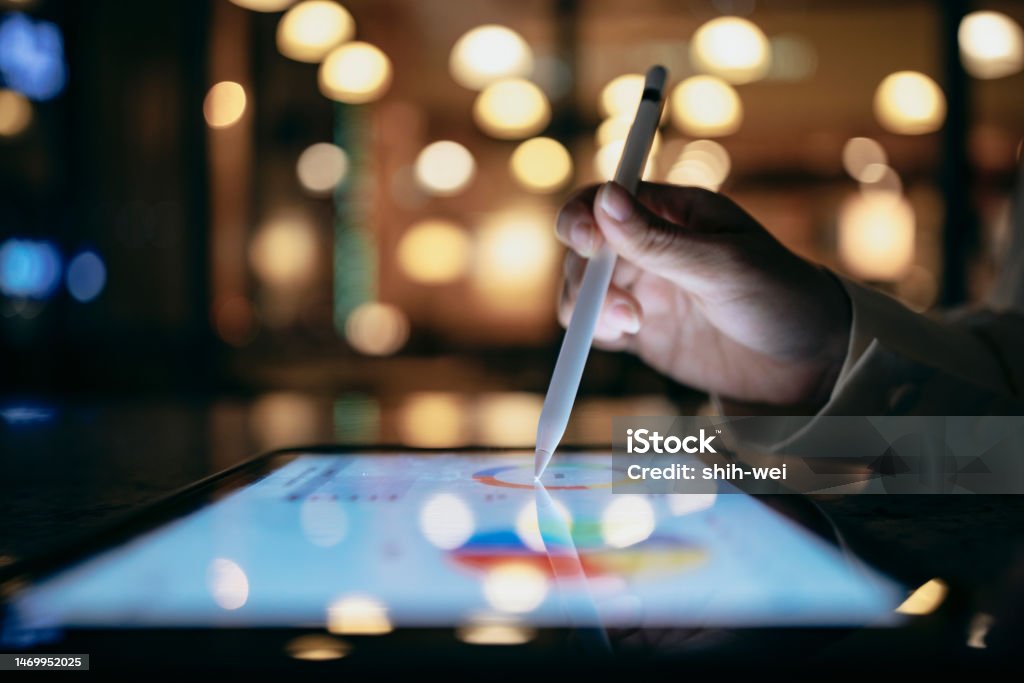 A business person is working in a cafe at night, using a tablet device and a stylus to look up chart indexes and using them for work projects. A businessperson is using a stylus to edit work reports on a tablet computer, which, combined with a mobile device, allows them to work more smoothly. Technology Stock Photo