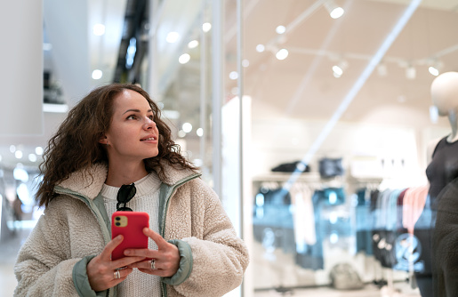 Young woman walking around the shopping mall looking at window of fashion stores and holding mobile phone in her hands.