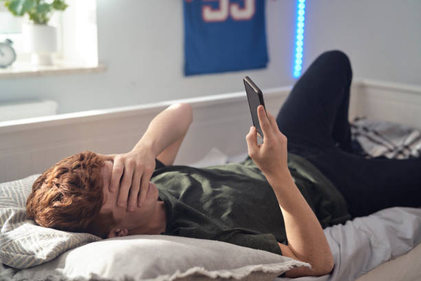 Shocked caucasian teenage boy covering eyes while browsing phone on bed stock photo