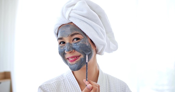 Asian woman applying grey mask on face smiling at camera. Spa beauty procedures at home for girl. Skincare, hydrating and moisturizing concept