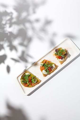 Aesthetic composition with salmon bruschetta on white background with shadows from flowers. Italian bruschetta with salmon, avocado, cheese and olives on fine dining in summer. Elegant menu concept