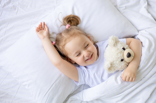 little cute blonde baby girl laughing in her sleep, sleeping on a white cotton bed at home hugging a teddy bear, close-up