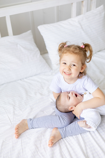 a little girl hugs a newborn sister or brother on a white bed at home, two children in the family
