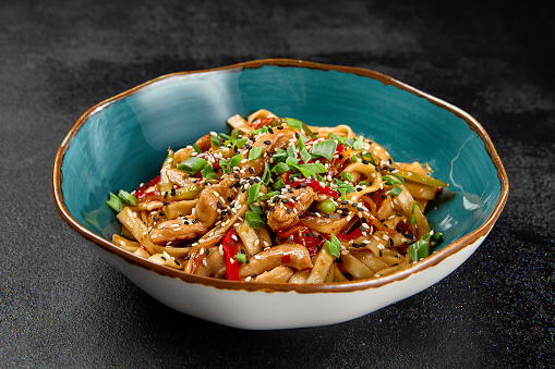 Traditional asian fried noodles with chicken and vegetables on dark background. Udon noodles with chicken on wok with sesame and green onion. Stir-fried udon with meat and vegetables on wok