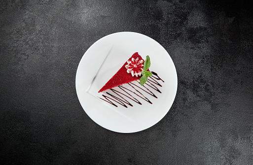 Red velvet cake on white plate on dark stone background. Popular red cake with layer biscuit and cream. Red velvet dessert in minimal style on black backdrop. Piece of cake with mint