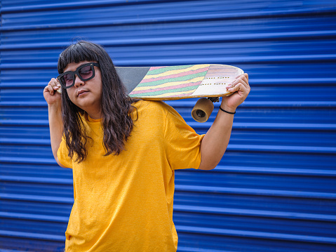 Portrait of long-haired asian skateboarder man wearing sunglasses and yellow t-shirt while holding skateboard on shoulder standing at outdoors blue wall background, looking at camera