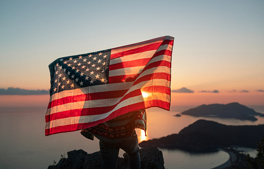 Proud woman raising the flag of the United States of America standing on the top of the mountain at sunset