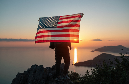 Proud man raising the flag of the United States of America standing on the top of the mountain at sunset