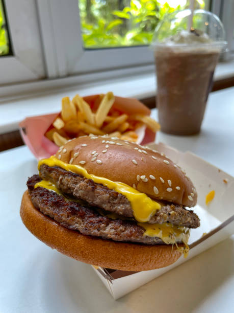 Stock photo showing close-up view of cheeseburger served in sesame seed burger bread bun in cardboard takeaway box. imagen de comida rápida para llevar hamburguesa doble con queso con pan de hamburguesa de semillas de sésamo en una caja de cartón en la mesa del restaurante, queso derretido, porción de papas fritas, batido de chocolate, enfoque en primer plano