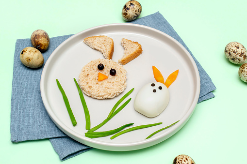 Easter funny creative healthy breakfast lunch food idea for kids, children.Bunny, rabbit made from boiled chicken eggs,bread, peeled carrots, greens on plate green table background.Top view Flat lay.