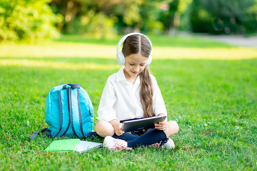 a first-grader girl with a backpack and a tablet with headphones on a green lawn reads a book or does homework, goes back to school
