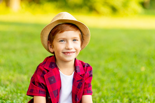 portrait of a little red-haired boy on a green lawn in a hat and a red shirt smiling cheerfully in summer