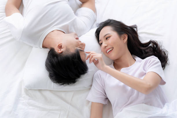 Happy Asian couple lying down smiling and relaxing together in bed. Happy Asian couple lying down smiling and relaxing together in bed. asian cuddling in bed stock pictures, royalty-free photos & images