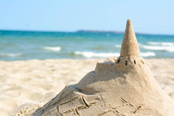 Stock photo showing close-up view of a sandcastle built on a sunny, golden sandy beach with sea at low tide in the background. Summer holiday, tourism and activities concept.