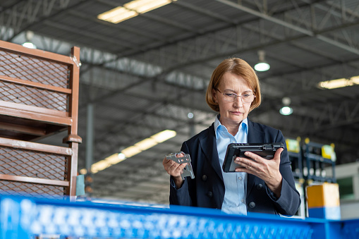 Product inspector and quality control improve sell performance and growth your business. Senior Caucasian warehouse manager woman holding spare part and inspector quality over tablet in the automative warehouse factory.