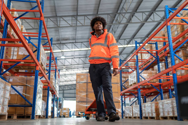 istock Reliability on-time in inventory management systems help businesses grow and be trusted. Front view of African American in reflective safety suit warehouse worker walking and drag hand lift pallet from distribution warehouse factory 1469984048