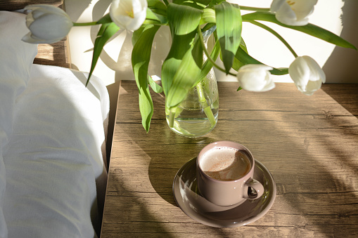 Beautiful white tulip bouquet and cup of coffee on nightstand in bedroom