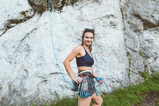 Part of young woman wearing safety harness, carabiners, shorts, standing in front of rock and smiling at camera.