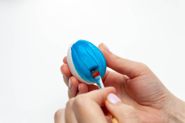 istock The hand holds a brush and paints an egg. The process of dyeing an Easter egg light blue. Brush in a woman's hand on a white background 1469989700