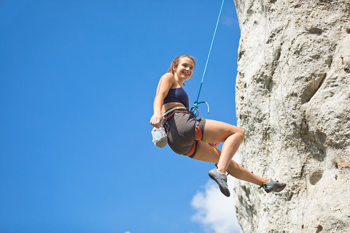 Young woman climbing on cliff rock with blue sky in the background, hanging on rope and smiling at camera.