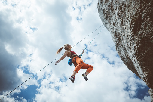 Wide angle view of young woman climbing on cliff rock with cloudy sky in the background, hanging on rope.