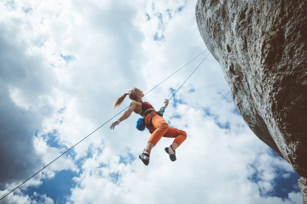 Young woman hanging on rope while climbing Wide angle view of young woman climbing on cliff rock with cloudy sky in the background, hanging on rope. adrenaline stock pictures, royalty-free photos & images
