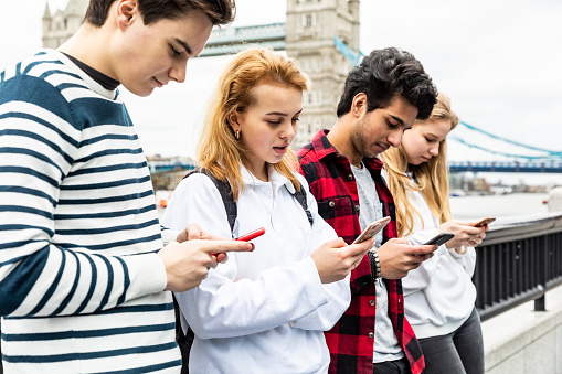 Group of teen students in London using phone near Tower Bridge - Multiracial group of teenagers best friends enjoying time together in London - Lifestyle and friendship in London