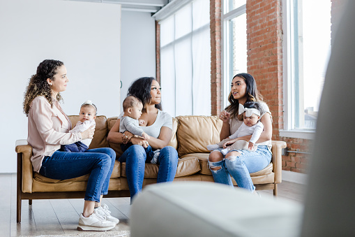 The diverse group of women sit on the couch and talk to each other about their mental health struggles as they adjust to being stay-at-home moms.