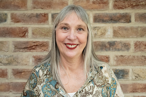 Front view of Caucasian woman with long grey hair and bangs, wearing casual blouse and red lipstick, standing against indoor brick wall and smiling at camera.