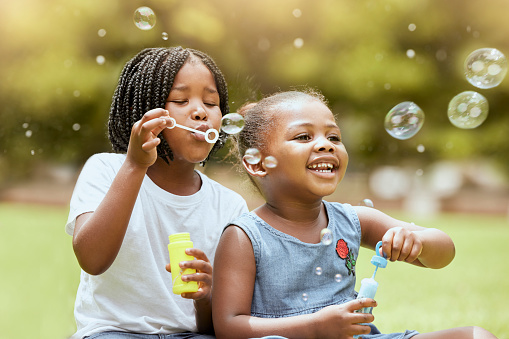 Black kids, children and blowing bubbles at park, having fun and bonding. Girls, happy sisters and playing with soap bubble toys, relax and enjoying quality time together outdoors in nature on grass