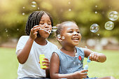 Black kids, children and blowing bubbles at park, having fun and bonding. Girls, happy sisters and playing with soap bubble toys, relax and enjoying garden together outdoors in nature on grass