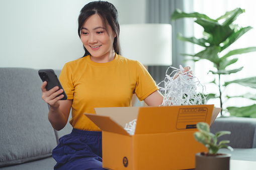 Asian woman unpacking carton box happy excited and using smartphone, sitting on sofa in living room at home. Happy woman enjoying online shopping surprised with fast delivery service.