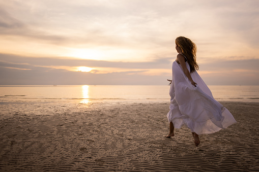 Stunning woman in white dress walking on the beach by the sea in sunset.