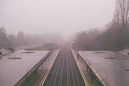 An outdoor metro station in the fog in Harrow, London - England. The railway tracks are centred in the image and leading towards a railway siding in the distance, where a train sits stationary.\n\nShot on CineStill 400D 35mm film.