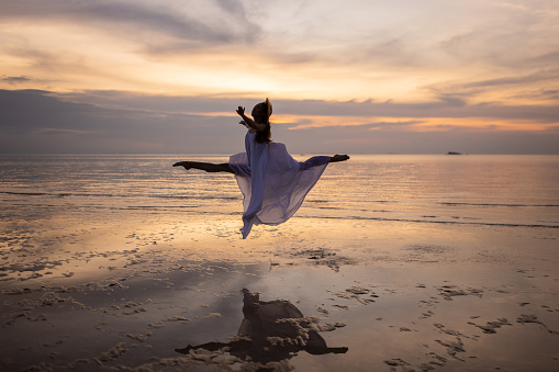 Stunning woman ballerina in white dress dancing on the beach by the sea in sunset.