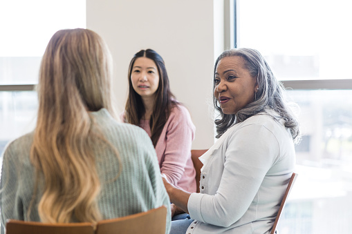 In group therapy, an unrecognizable young adult woman listens to advice from the female senior adult counselor as the mid adult woman looks on.