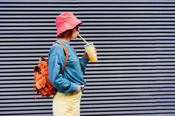 Stylish girl in bright clothes and bucket hat drinking sugar flavored tapioca bubble tea while walking near gray striped urban wall. Portrait of fashionable hipster girl. Street fashion. Copy space. Stylish girl in bright clothes and bucket hat drinking sugar flavored tapioca bubble tea while walking near gray striped urban wall. Portrait of fashionable hipster girl. Street fashion. Copy space bucket hat stock pictures, royalty-free photos & images