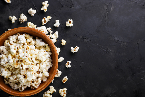 Popcorn in a wooden plate and scattered on a dark background, space for text. Top view