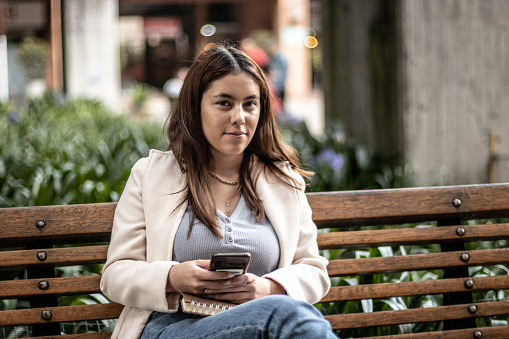 Portrait of a young student sitting on bench while using mobile phone outdoors