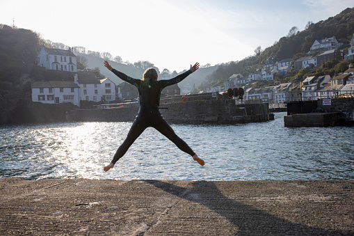 Woman jumping into the sea while on a staycation in Polperro, Cornwall. She is wearing a wetsuit and has her arms in the air, excited about being on vacation.