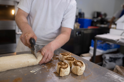 Medium shot of an unrecognisable bakery worker making a cinnamon pastry at his small bakery business in Polperro, Cornwall. He is sectioning the roll into pieces.